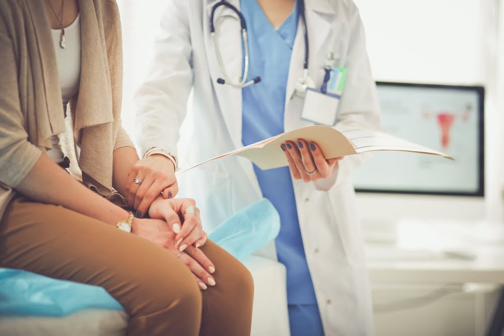 imagine cu un medic si un pacient. Medicul îl ține de mana pe pacientul așezat pe scaun, iar in cealalta mana ține o listă pe care se află suplimente cu fier pentru adulți